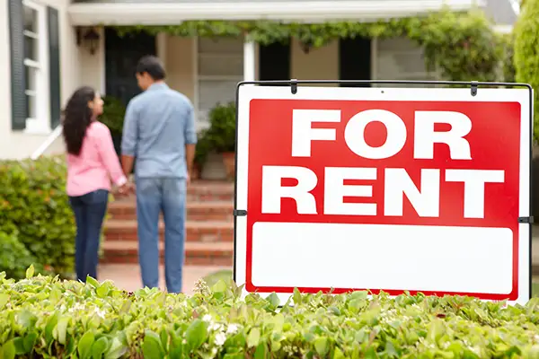 A red “for rent” sign in front of a rental property. In the background, two potential tenants can be seen viewing the property. Property management companies can make it easier for landlords to rent out their homes.